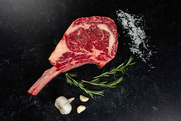 Certified ONYA® Tomahawk Ribeye photographed raw with beautiful marbling. Shown with garnishes including sea salt, rosemary, and whole garlic cloves. 