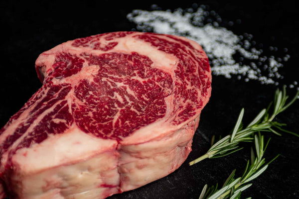 Certified ONYA® Tomahawk Ribeye photographed raw with beautiful marbling. Shown with garnishes including sea salt and rosemary. Incredibly tender steak