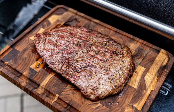 A reverse seared Certified ONYA® beef flank steak sits on a wooden cutting board on the prep tray of a Pit Boss pellet smoker grill. 
