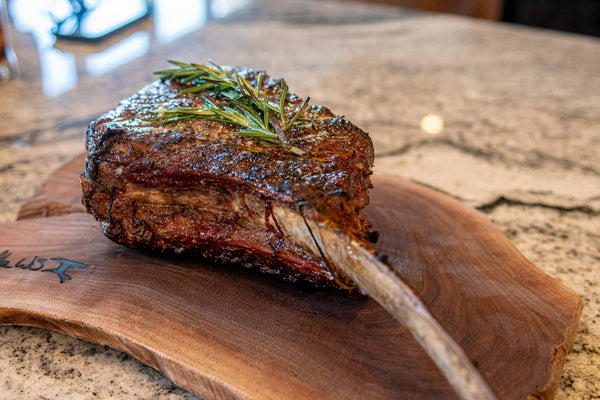 Large Certified ONYA® Tomahawk Ribeye 3 inches thick shown cooked on a wooden cutting board on a marble countertop. Rosemary can bee seen resting on top of the perfectly cooked steak