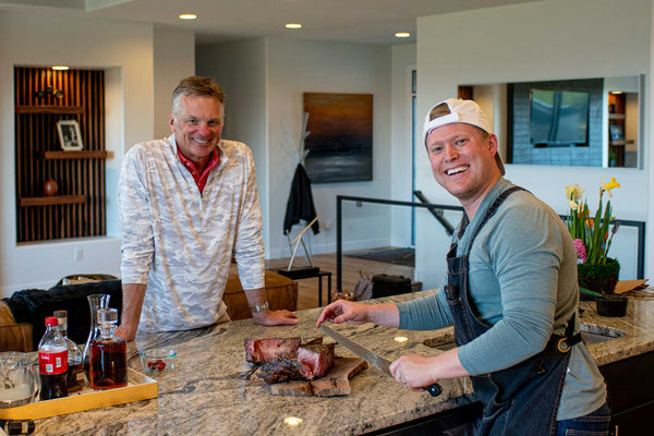 Two friends shown slicing a Certified ONYA® Tomahawk Ribeye steak from BetterFed Beef. Pictured standing around a marble kitchen island
