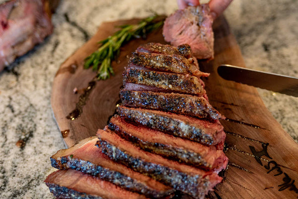 3.75lb Certified ONYA® Tomahawk Ribeye Steak cooked to medium rare with the reverse sear method on a charcoal grill and sliced for serving on a wooden cutting board. Rosemary can be seen on top of the as tender as Wagyu steak while it rests before serving alongside the bone.
