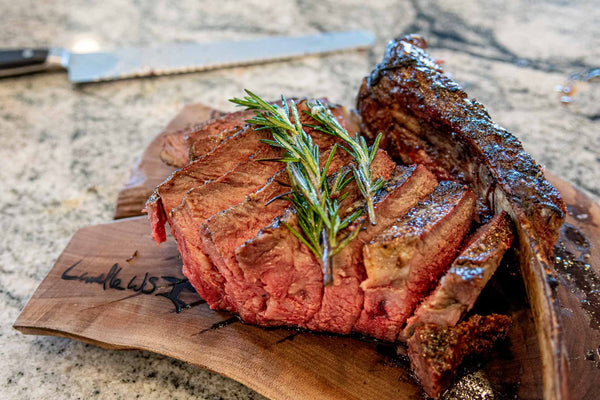 60oz Certified ONYA® Tomahawk Ribeye Steak cooked to medium rare with the reverse sear method on a charcoal grill and sliced for serving on a wooden cutting board. Rosemary can be seen on top of the as tender as Wagyu steak while it rests before serving alongside the bone.  