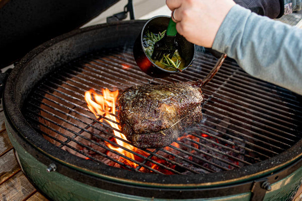 60 ounce BetterFed Beef Certified ONYA® Tomahawk Ribeye being seared over an open flame on a Big Green Egg charcoal grill. The chef is basting the incredibly tender steak with butter and rosemary for added flavor and to create a beautiful crust on this medium rare piece of beef. 