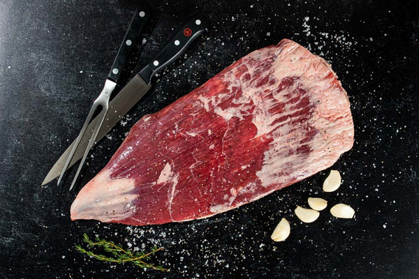 A certified ONYA® beef flank steak from BetterFed Beef with a kitchen chefs knife and a carving fork. Seasoned with salt and pepper, this makes one of the best steaks online you can purchase and have delivered to you home.